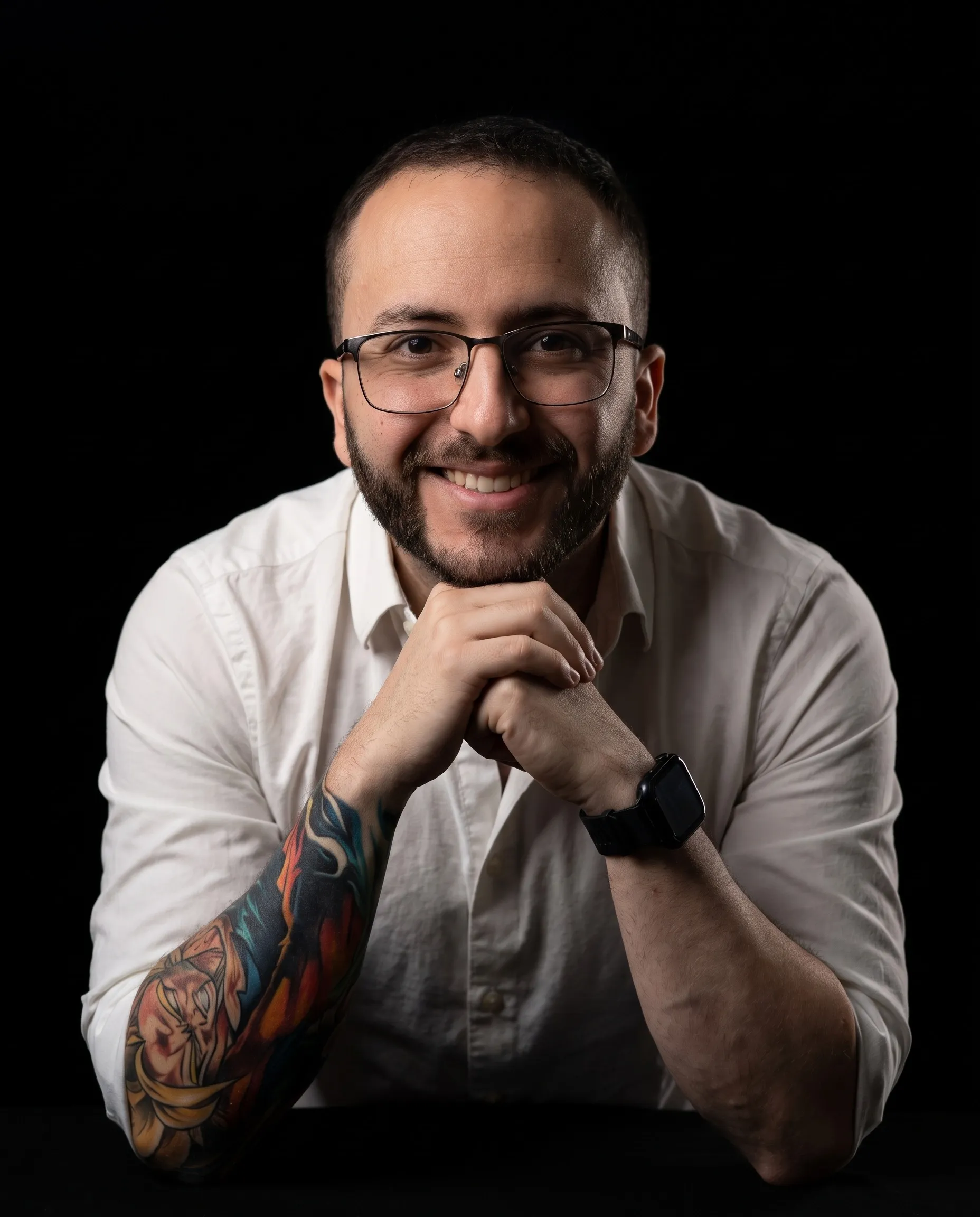 Andres Clavijo smiling at the camera in glasses and a white shirt, leaning on his forearms with his hands clasped under his chin, a colourful tattoo sleeve on one arm, lit from the side against a black background.